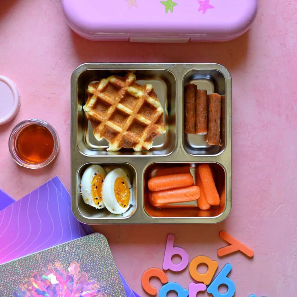Brunch-inspired school lunch with a Belgian waffle, hard-boiled egg, baby carrots and maple chicken stick.