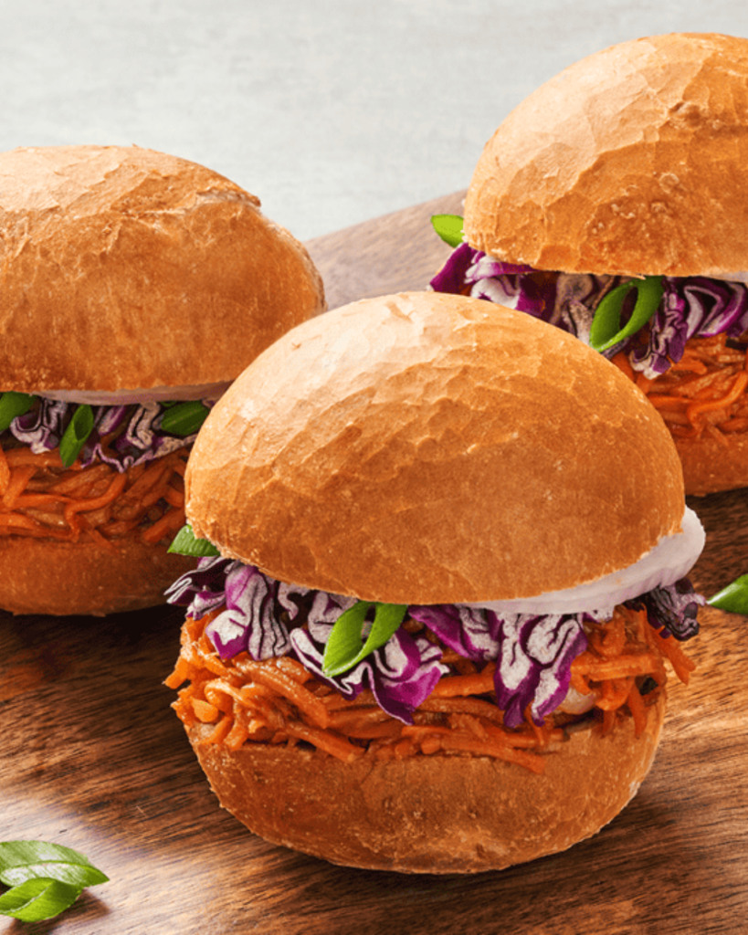 Three pulled BBQ carrot sliders topped with shredded purple cabbage, sliced onion and green onions on a wooden board.